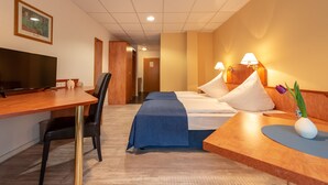 A hotel room with a wooden desk, a bed with a blue bedspread, two bedside lamps, a TV, and a framed picture on the wall. - Boardinghouse Burgpark (Huerth)