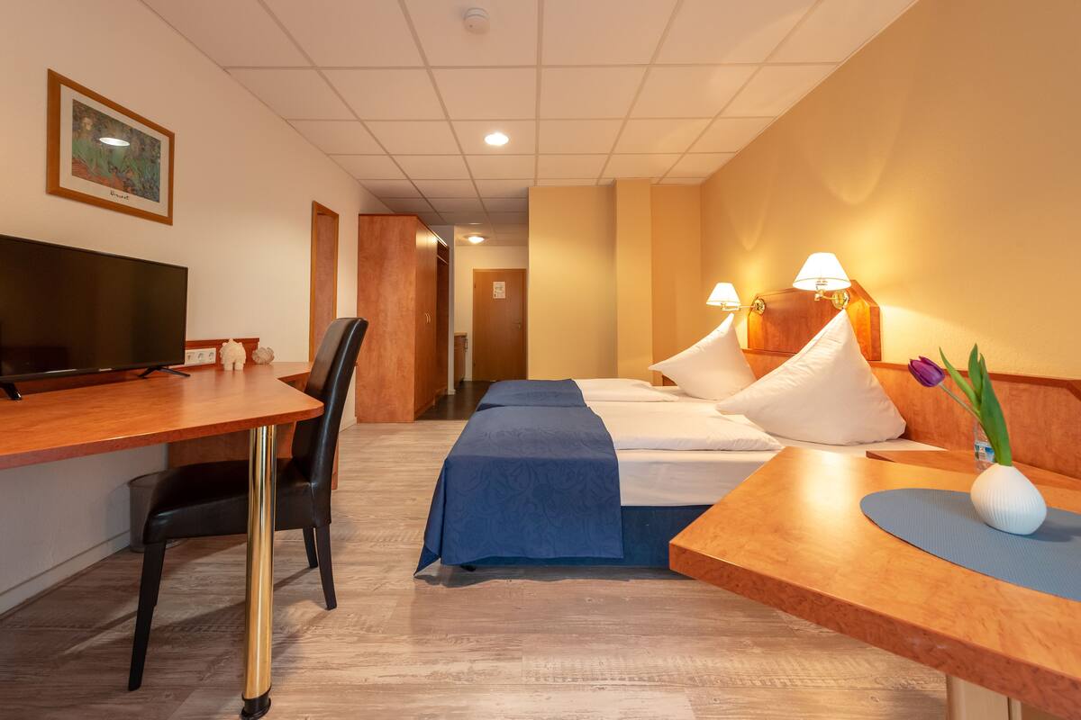 A hotel room with a wooden desk, a bed with a blue bedspread, two bedside lamps, a TV, and a framed picture on the wall.