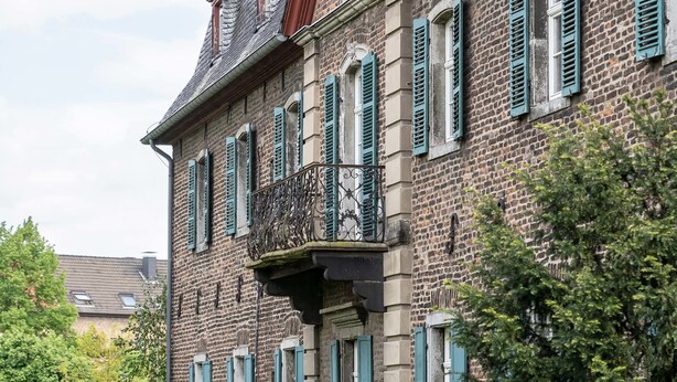 A two-story brick building with green shutters, a balcony with wrought iron railings, and a statue on the roof. - Boardinghouse Burgpark (Huerth)