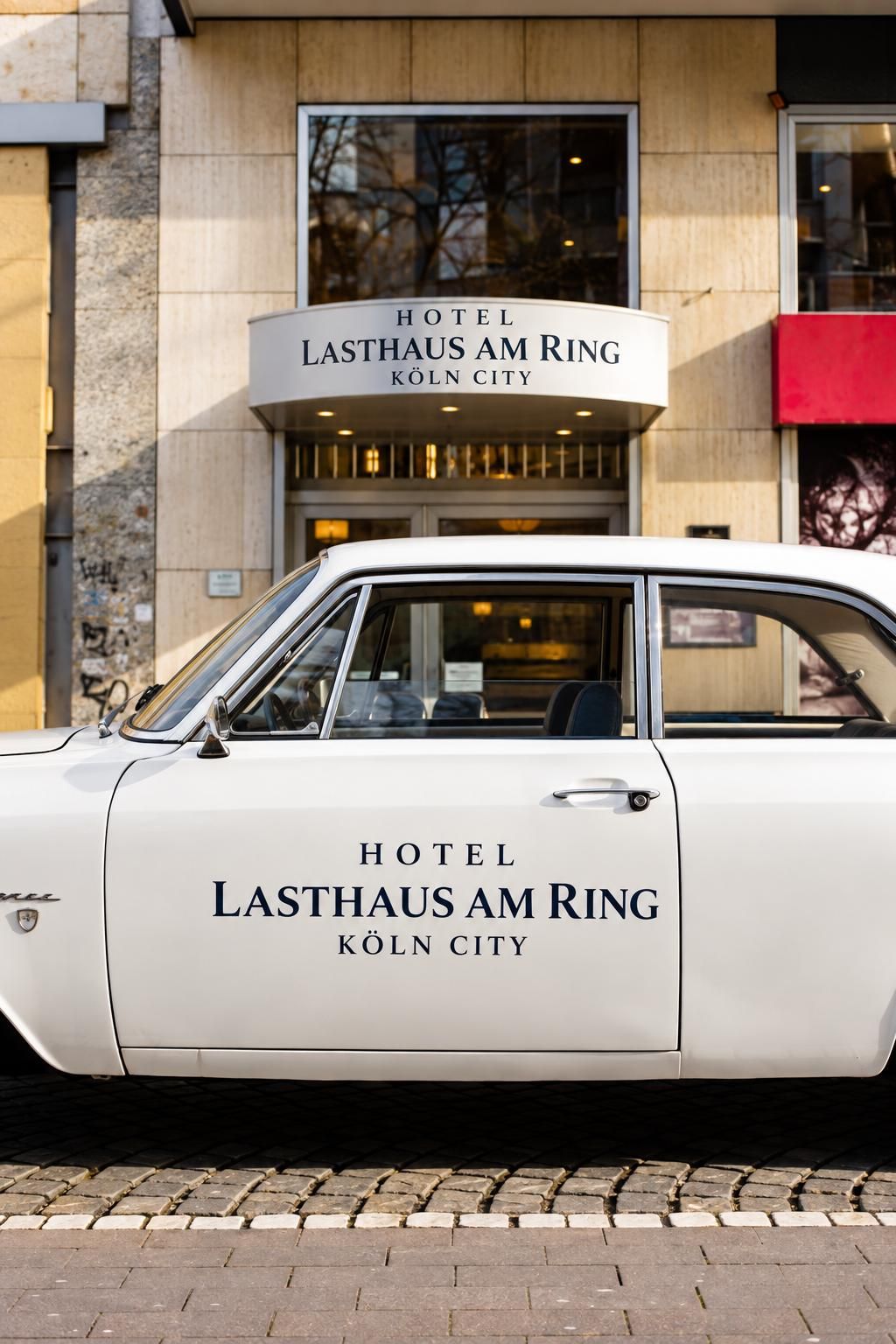 A white car parked in front of a hotel with the name 'HOTEL LASTHAUS AM RING KÖLN CITY' on its side.