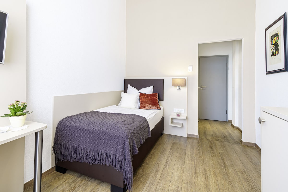 Une chambre à coucher moderne avec un lit, des tables de chevet, une petite table, une lampe fixée au mur et une plante en pot.