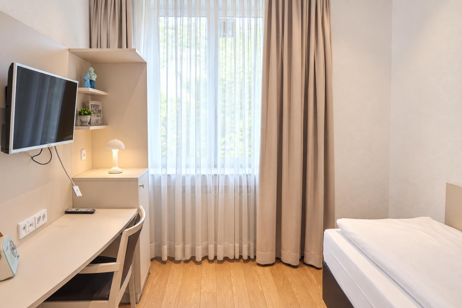 A hotel room with a bed, a desk with a TV, a lamp, and a window with curtains.