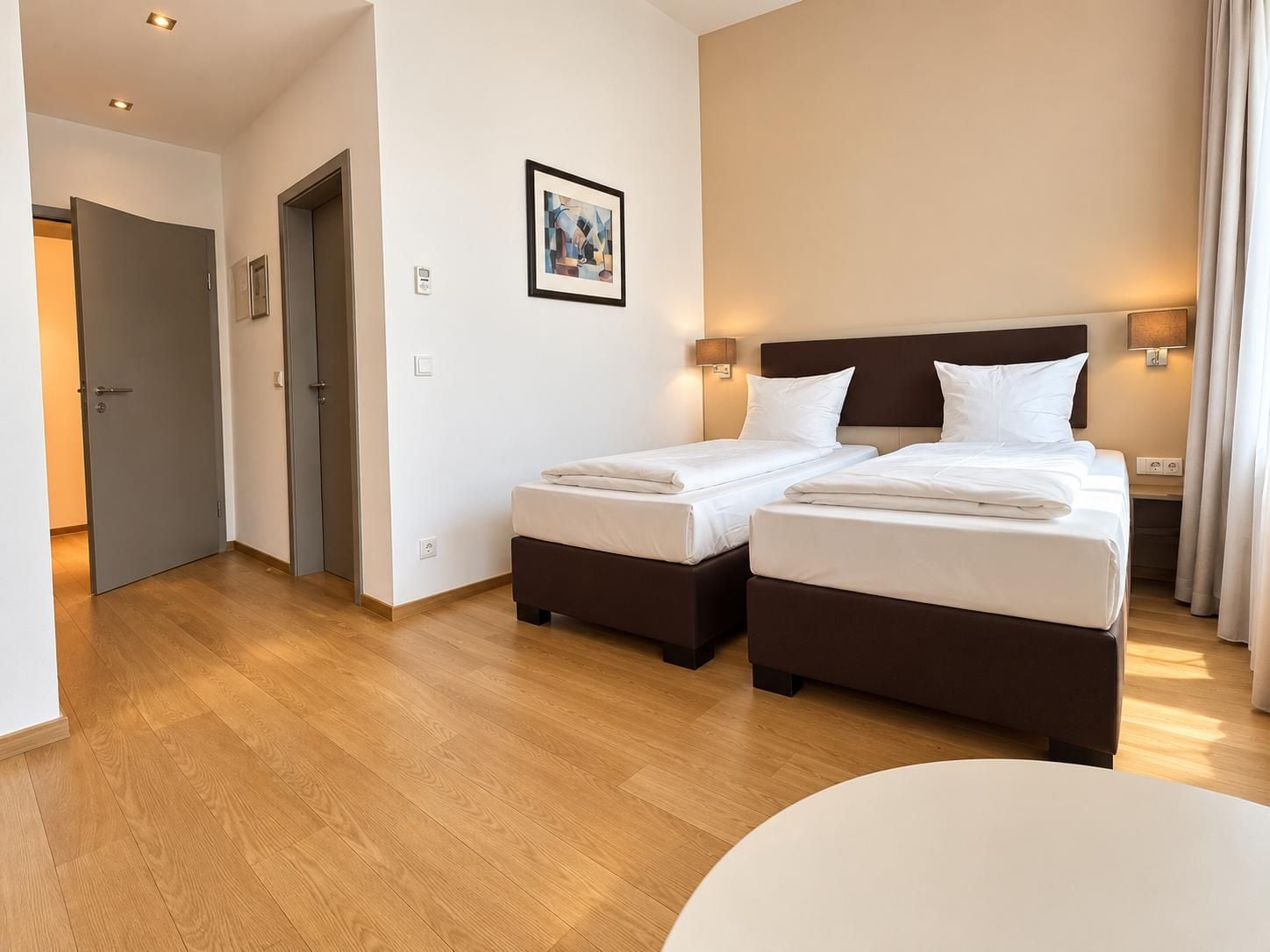 Une chambre d’hôtel avec deux lits, une table de chevet, une lampe fixée au mur et une porte donnant sur une autre pièce.