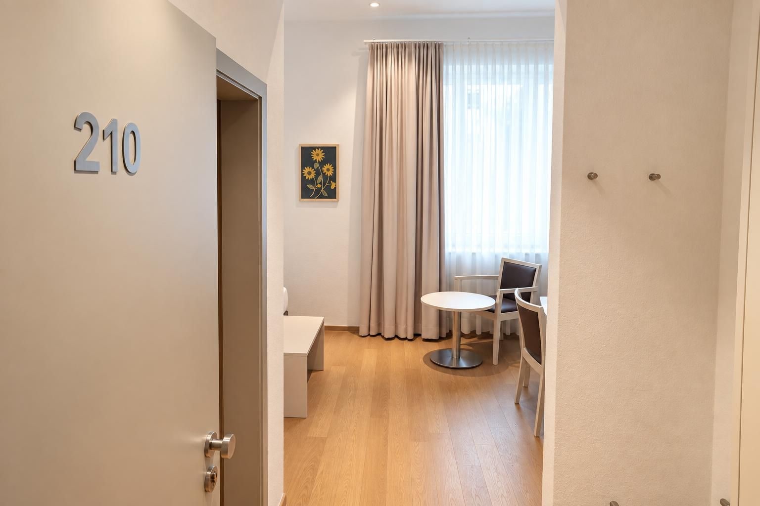 Une chambre d’hôtel avec une porte portant le numéro « 210 », une petite table avec deux chaises, un lit et une fenêtre avec des rideaux.