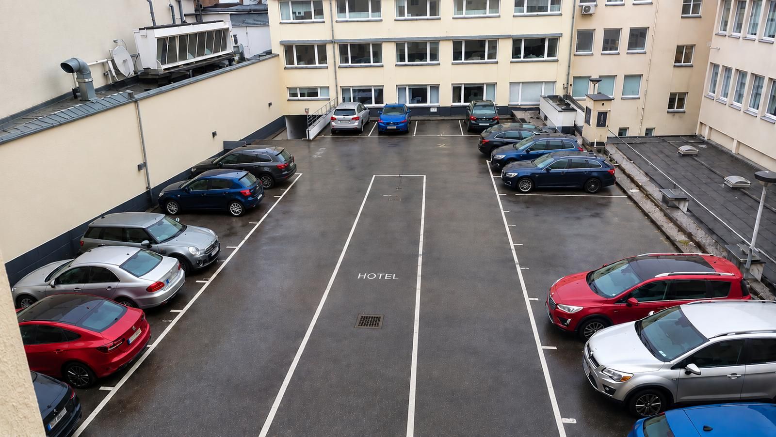 A multi-story hotel parking lot with several cars parked, a visible manhole cover, and a building with multiple windows and balconies.