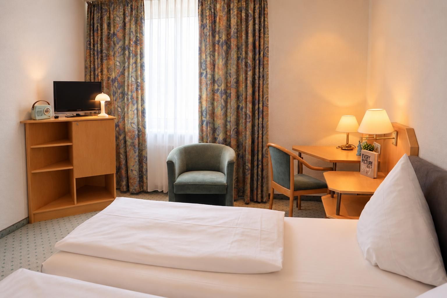 A hotel room with a bed, a desk with a lamp, a chair, a television, and a bookshelf.