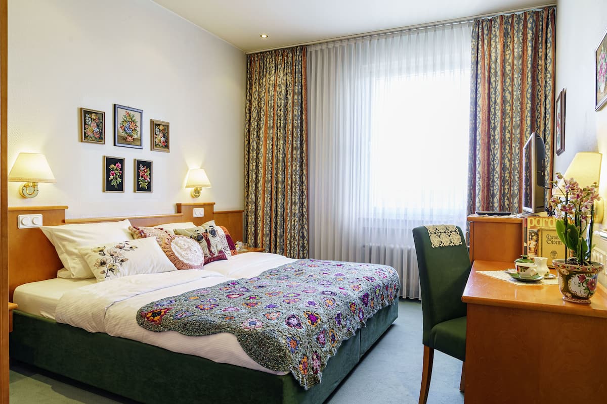 Une chambre d’hôtel avec un lit, deux lampes de chevet, un bureau avec une chaise, un vase de fleurs et des rideaux.
