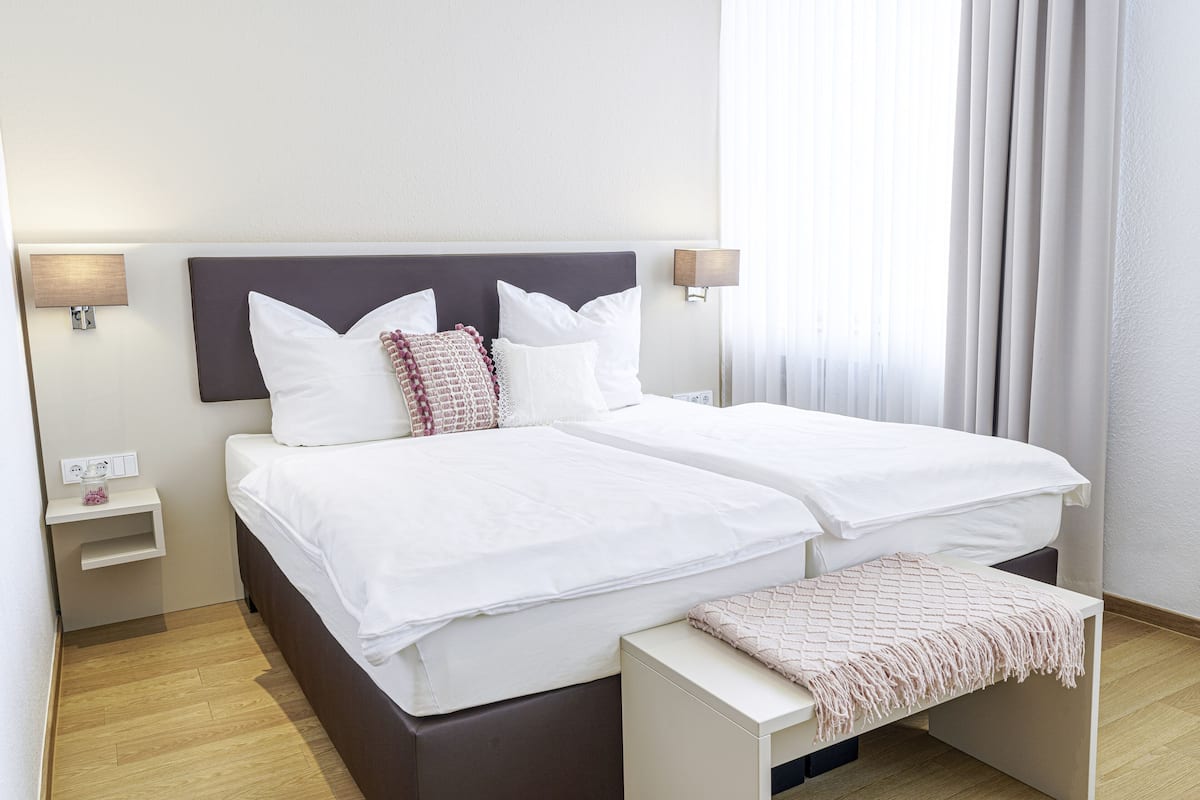 Un lit double avec du linge de lit blanc, une table de chevet avec une lampe et un banc agrémenté d’un plaid à motifs.