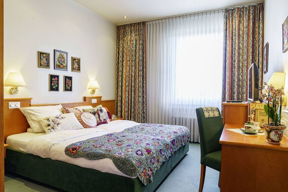 Une chambre d’hôtel avec un lit, deux lampes de chevet, un bureau avec une chaise, une plante en pot et une fenêtre avec des rideaux.