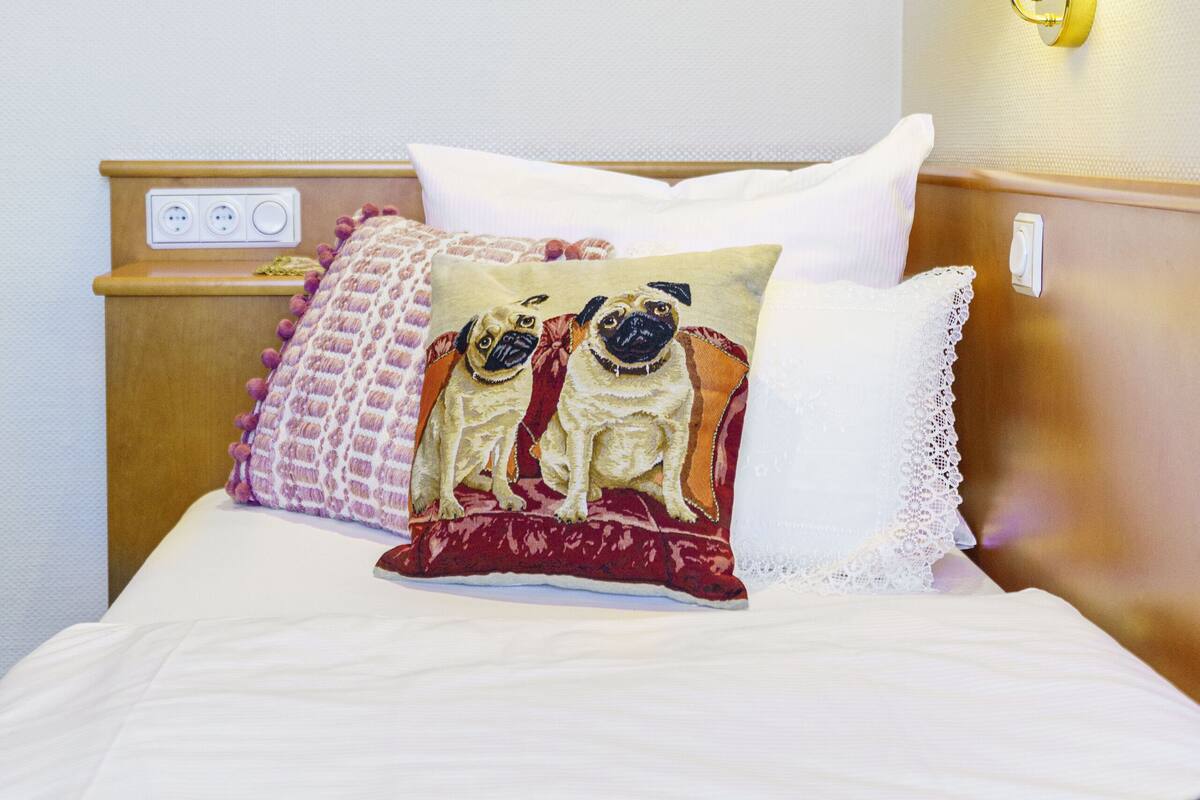Un lit bien fait, recouvert d’une couvre-lit à motifs, agrémenté d’un coussin décoratif orné d’un dessin de carlin, et une lampe fixée au mur.