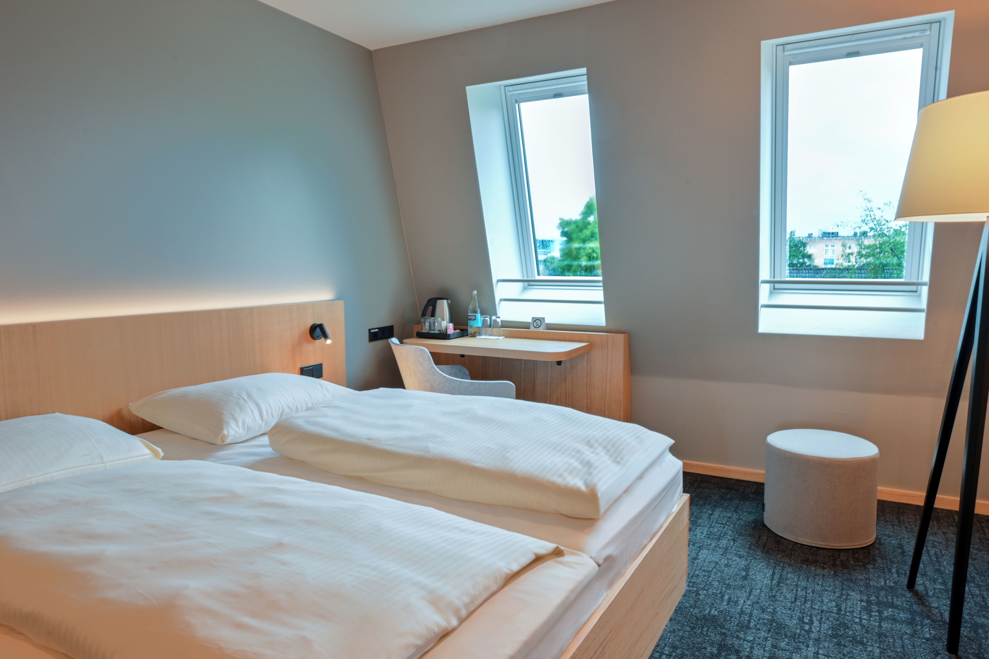 Habitación de hotel con dos camas, un escritorio, una silla, una lámpara y vistas de la ciudad a través de dos ventanas.
