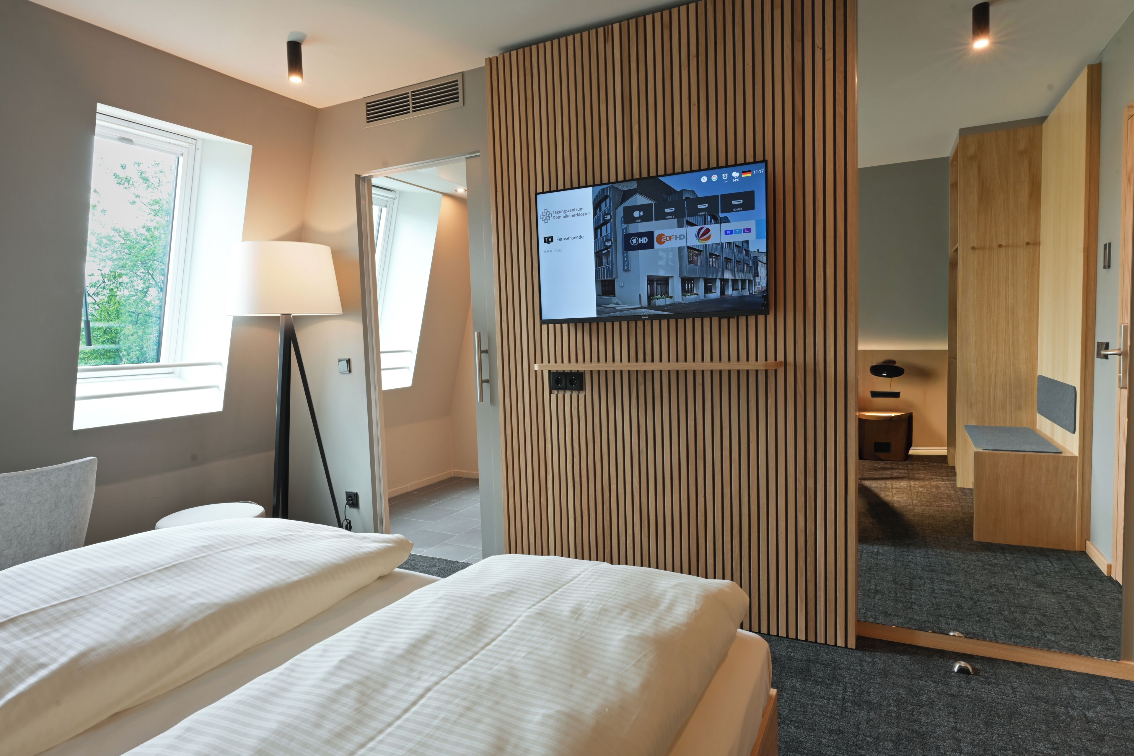 Een hotelkamer met een bed, een aan de muur bevestigde televisie, een lamp en een raam.