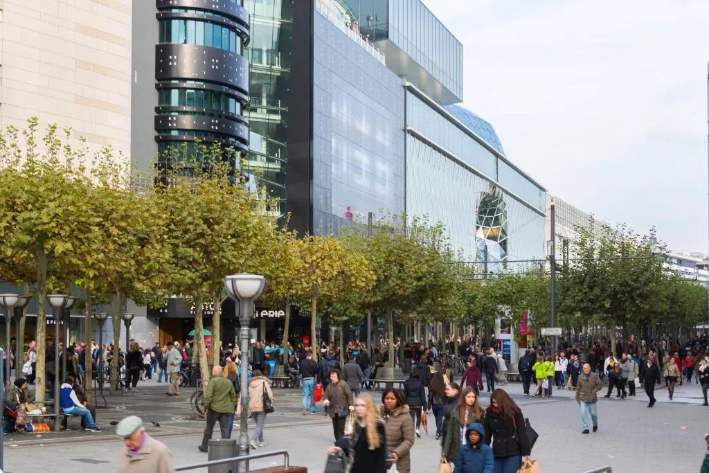 A busy urban street with pedestrians, trees, and modern buildings.