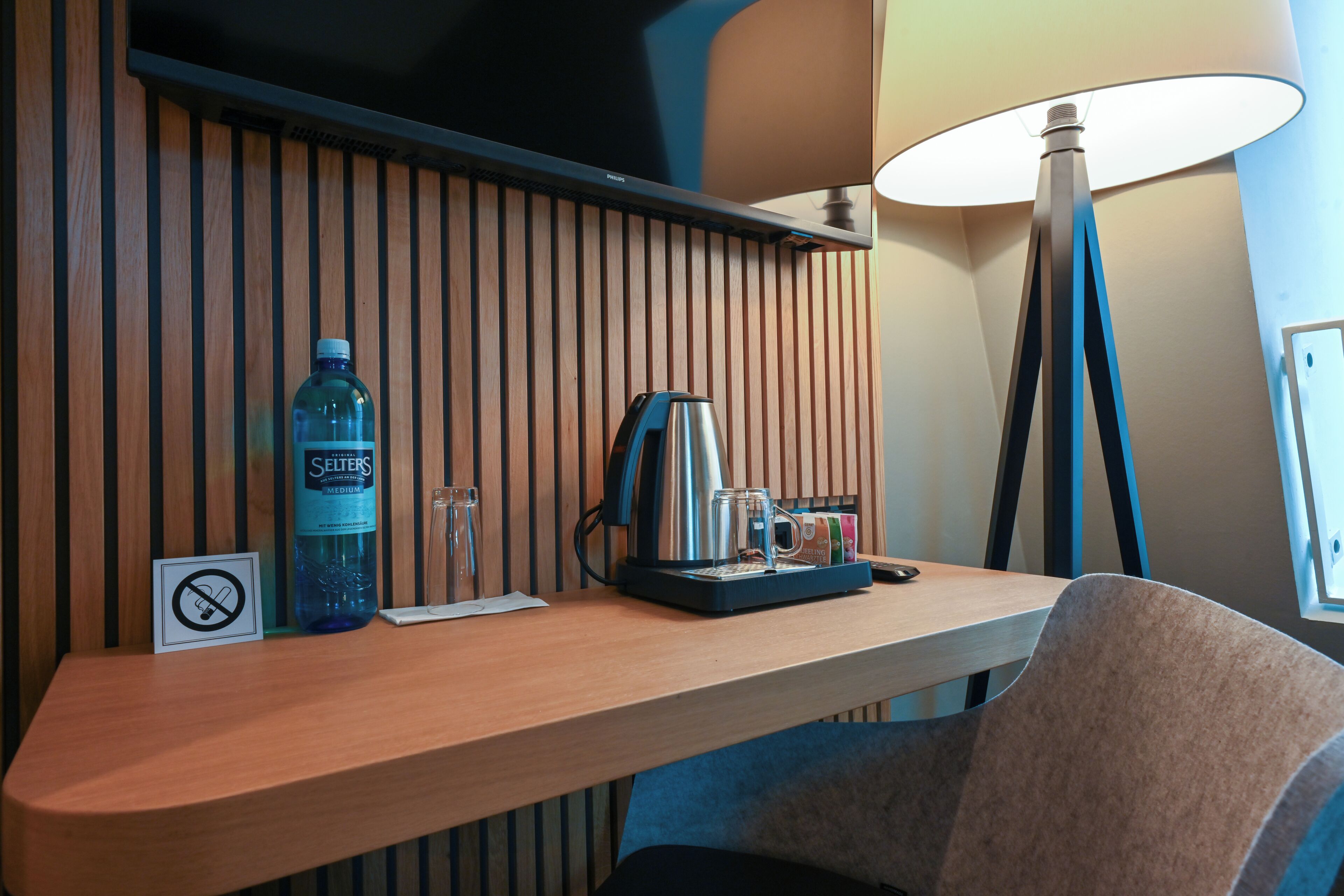 A hotel room with a wooden panel wall, a desk with a water bottle, a coffee maker, and a lamp.