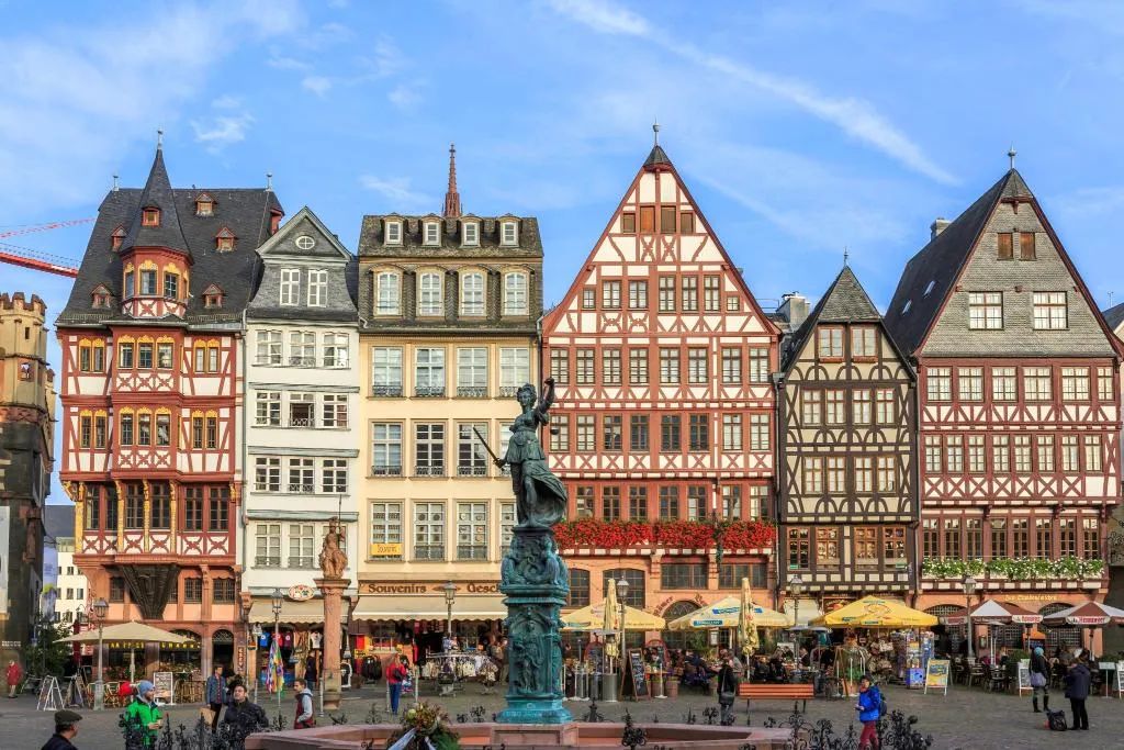 Ein historischer europäischer Stadtplatz mit traditionellen Fachwerkhäusern, einer zentralen Statue und Außenplätzen.
