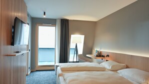 A hotel room with two beds, a desk, a chair, a TV, and a view of the sea.