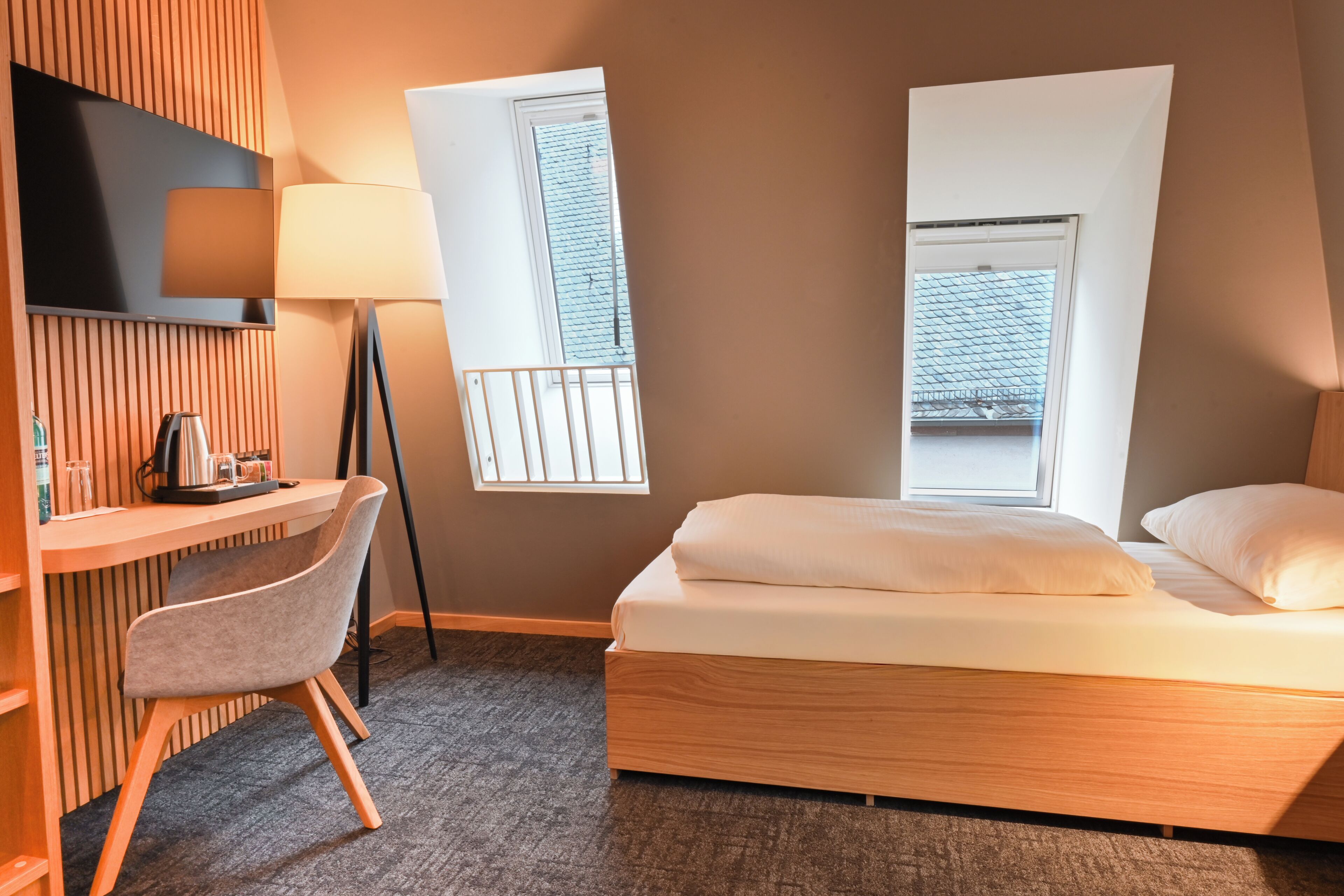 A hotel room with a bed, a desk, a chair, a TV, and two windows.