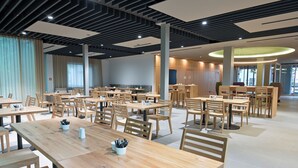 A spacious dining area with multiple wooden tables and chairs, a modern ceiling design, and large windows allowing natural light.