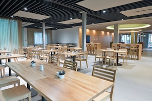 Un'ampia sala da pranzo con diversi tavoli e sedie in legno, un moderno design del soffitto e ampie finestre che lasciano entrare la luce naturale.