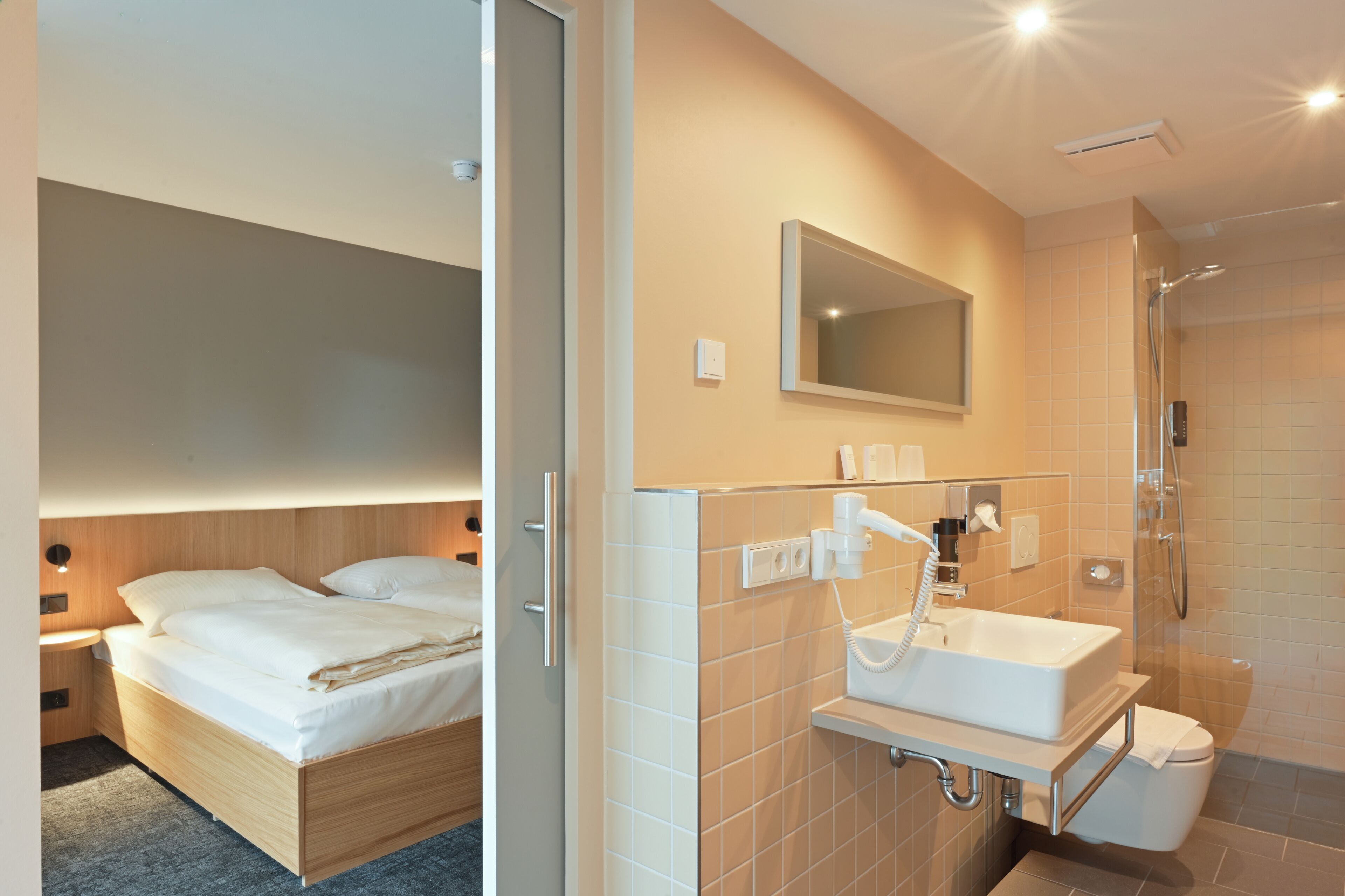A modern hotel room with a bed, a bathroom with a sink, mirror, and shower.