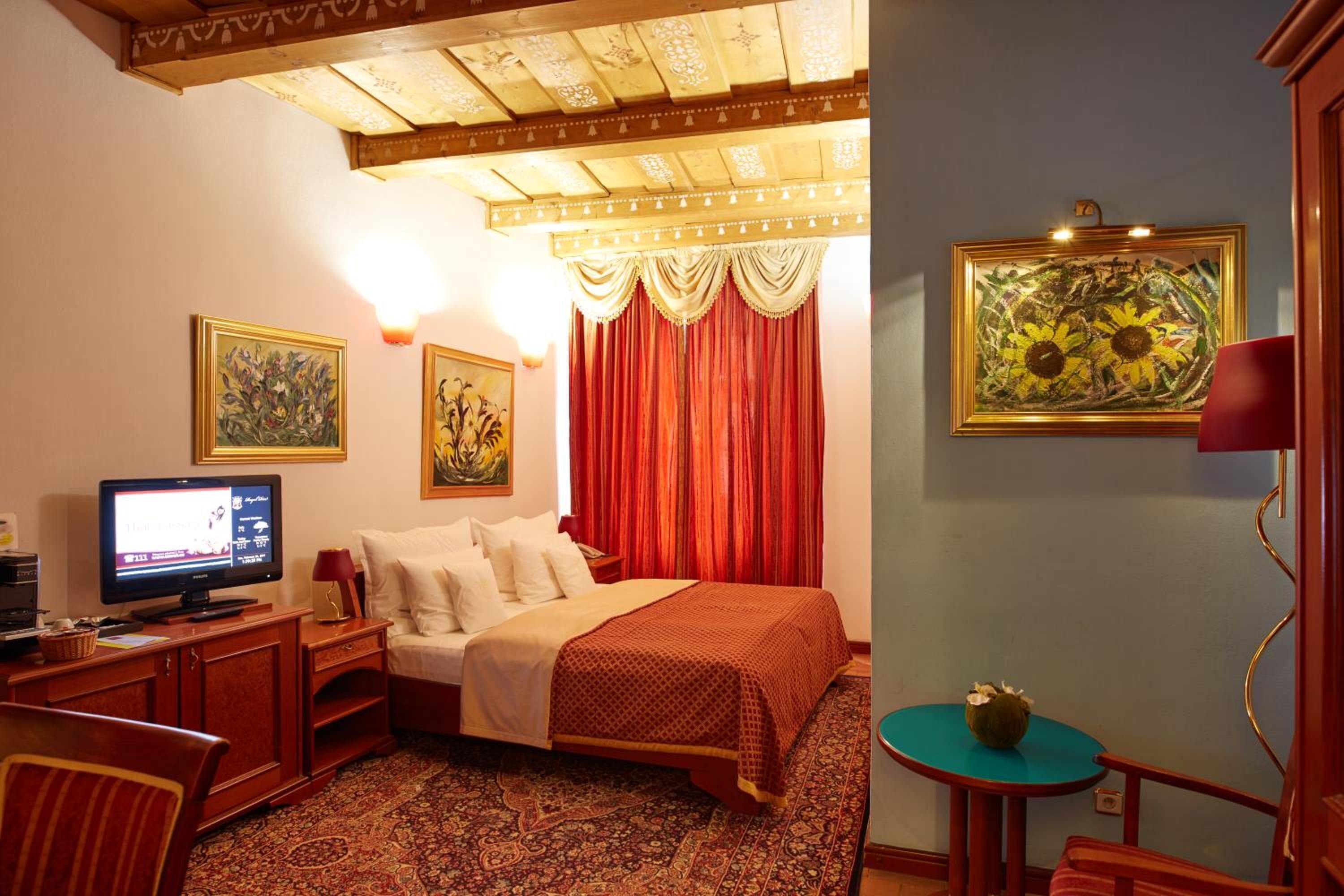 A hotel room with a bed, a desk with a computer, a television, a small table with a bowl, and two paintings on the wall.