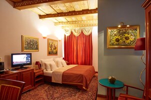 Una habitación de hotel con cama, escritorio con computadora, televisor, una mesita con un frutero y dos cuadros en la pared.