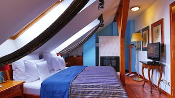 A bedroom with a sloped ceiling, a bed with white linens, a blue and gold striped bedspread, a wooden nightstand, a desk with a lamp, and a television on a stand.