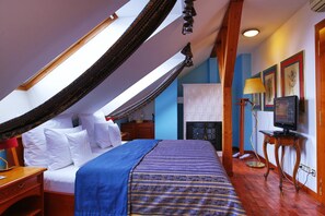 A bedroom with a sloped ceiling, a bed with white linens, a blue and gold striped bedspread, a wooden nightstand, a desk with a lamp, and a television on a stand.
