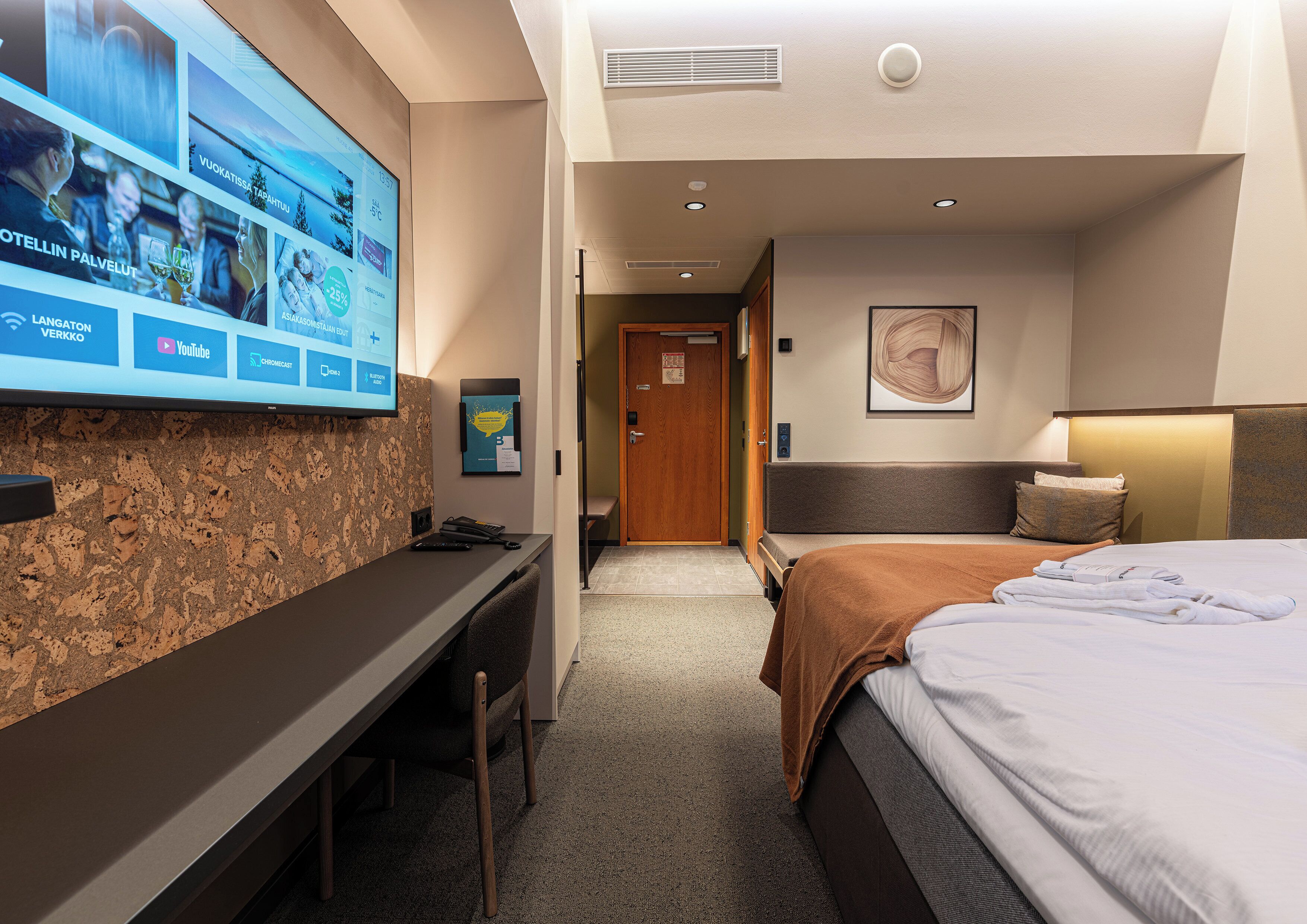 Una habitación de hotel con cama, escritorio, televisor mostrando un menú y puerta que da a otra habitación.