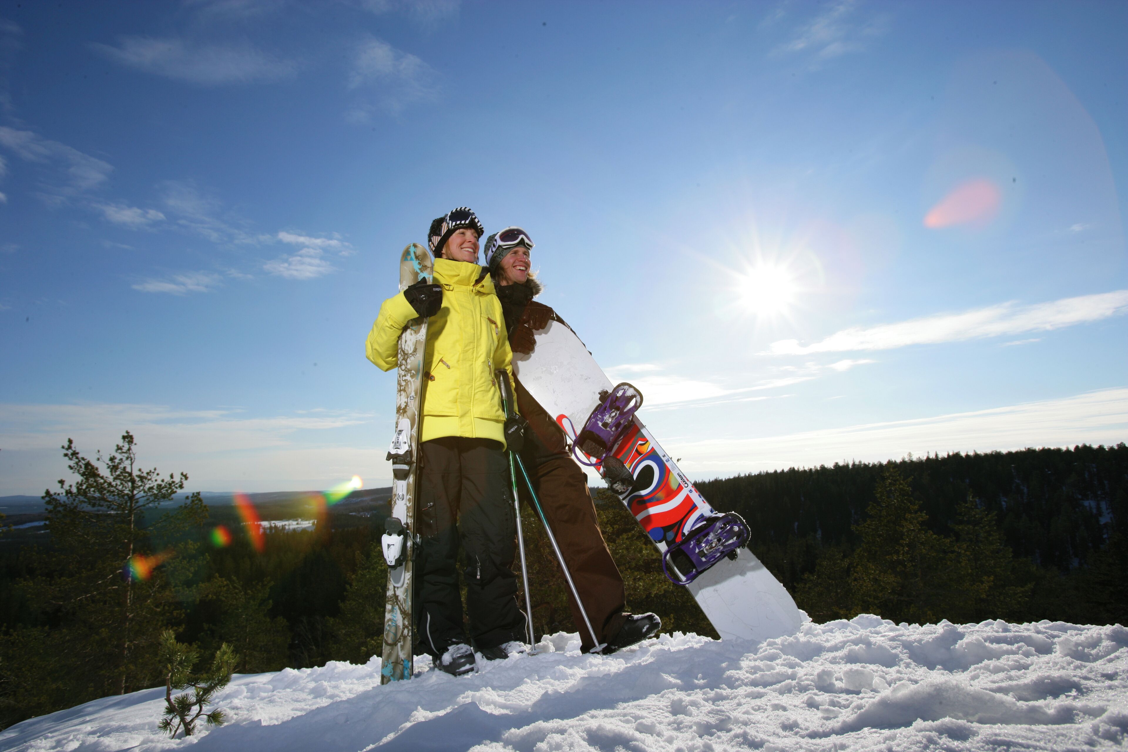 Duas pessoas em uma montanha nevada, com uma prancha de snowboard e esquis, com uma floresta e céu claro ao fundo.