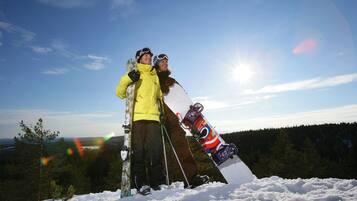 Duas pessoas em uma montanha nevada, com uma prancha de snowboard e esquis, com uma floresta e céu claro ao fundo.