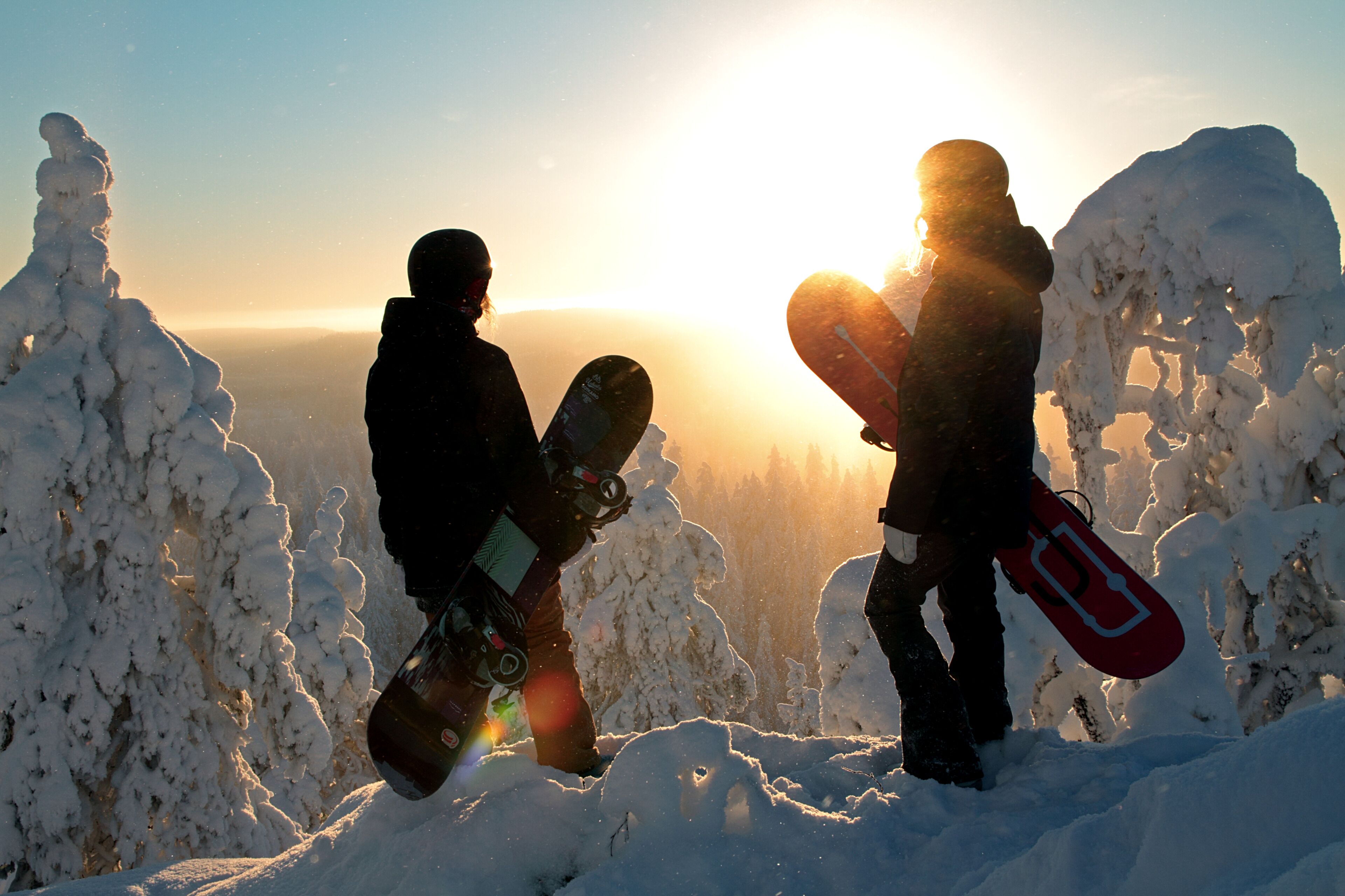 Two people holding snowboards on a snowy mountain with the sun in the background.