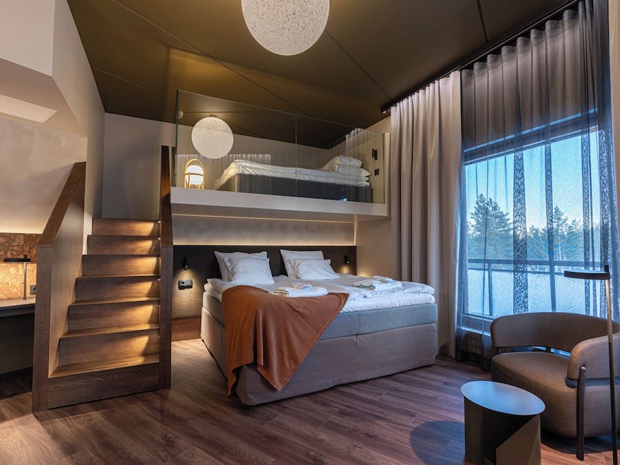 A modern hotel room with a bunk bed, a wooden staircase, a seating area, and a large window with a view of trees.