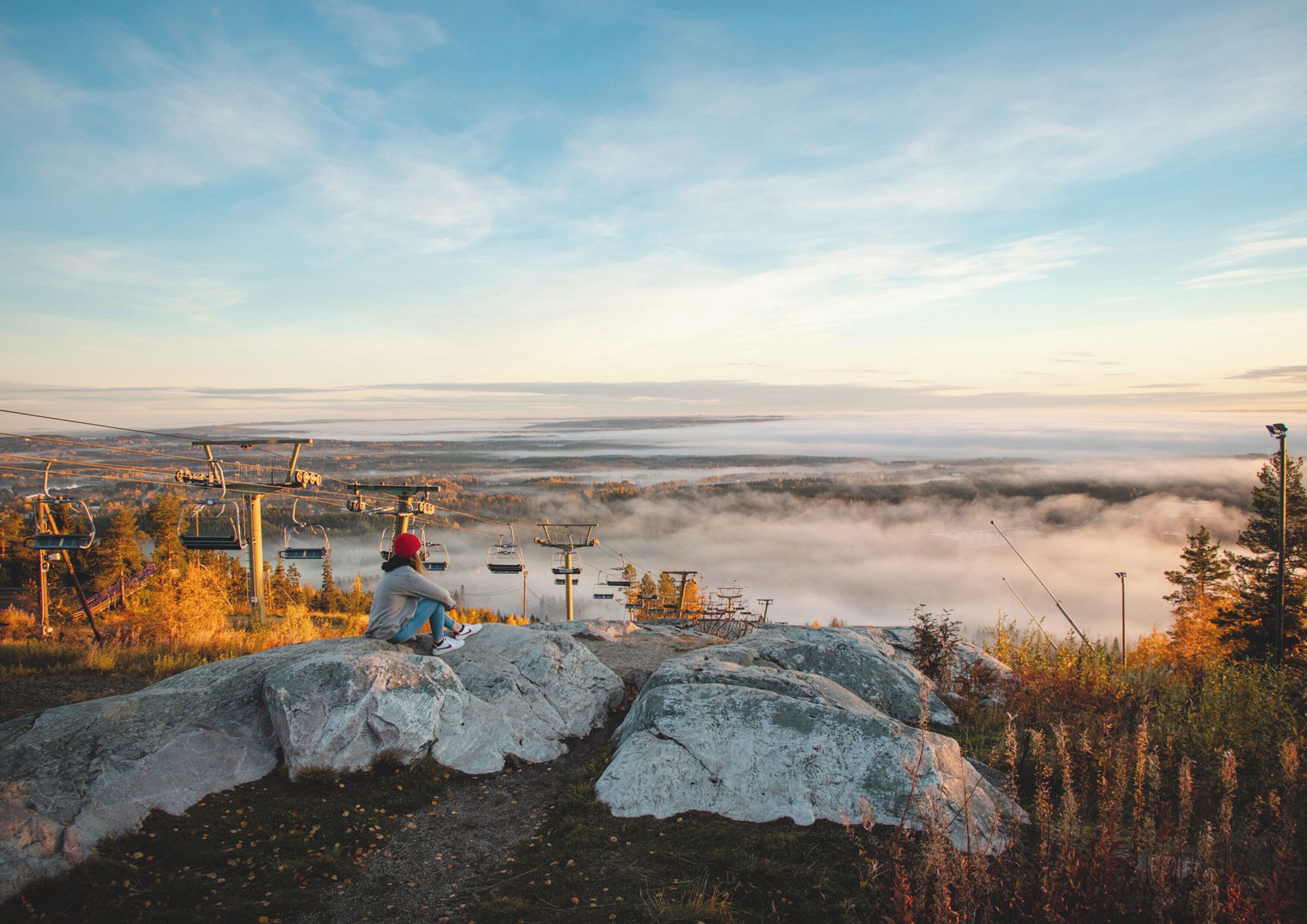 A person sitting on a rock with a view of a mountain range and a ski lift in the background.