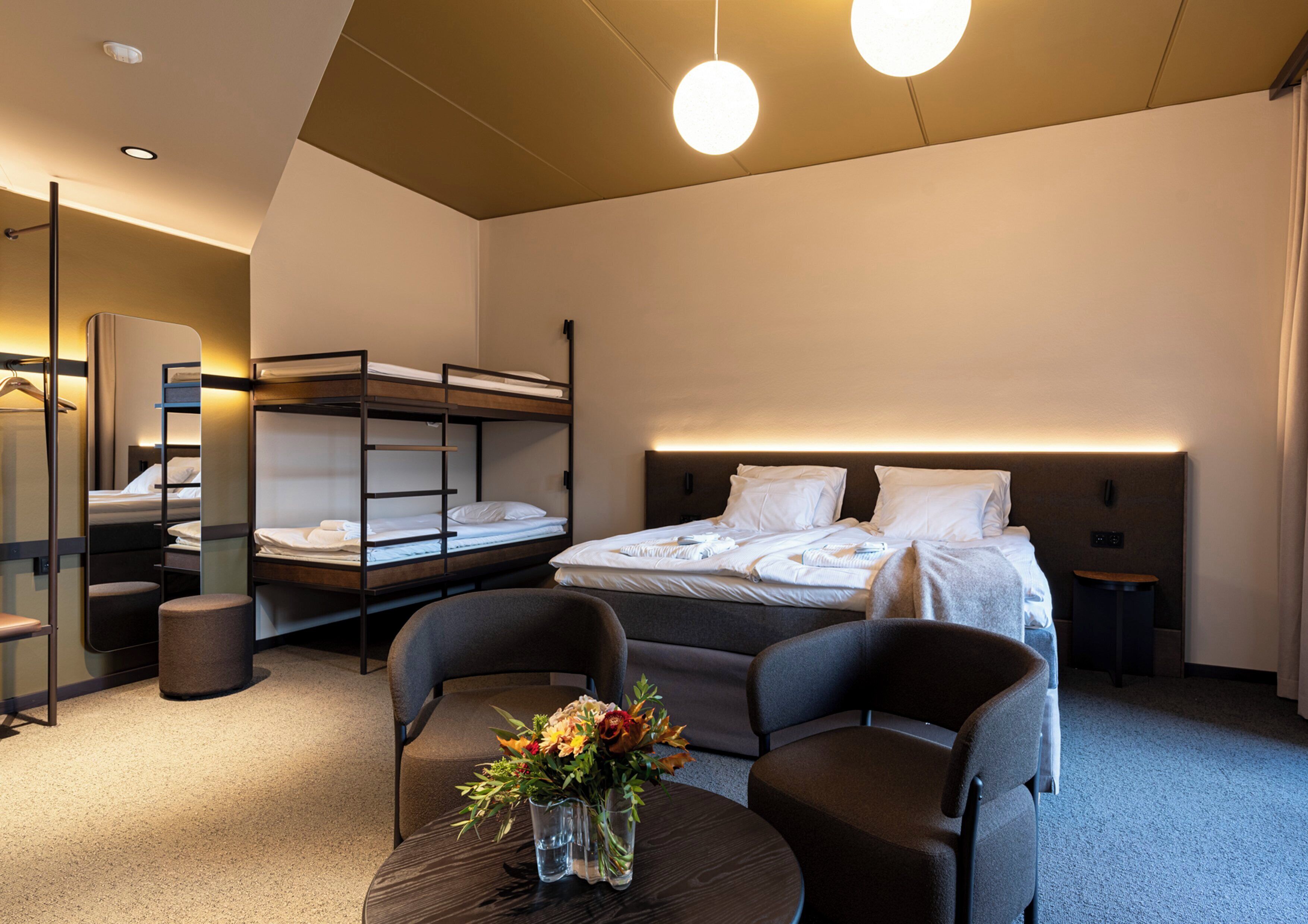 Habitación de hotel con literas, un escritorio con sillas, una mesita con un florero y un espejo.