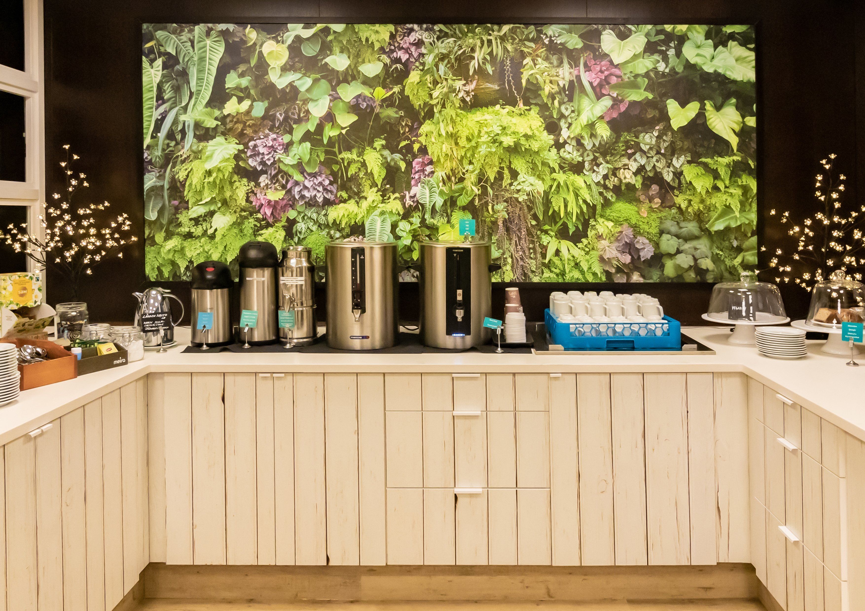 A coffee station with various coffee makers, a blue tray of cups, and a large wall mural of greenery.