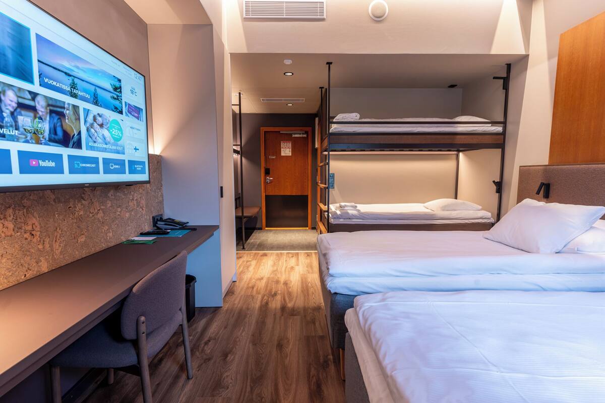 A hotel room with a desk, two beds, a TV mounted on the wall, and a wooden floor.