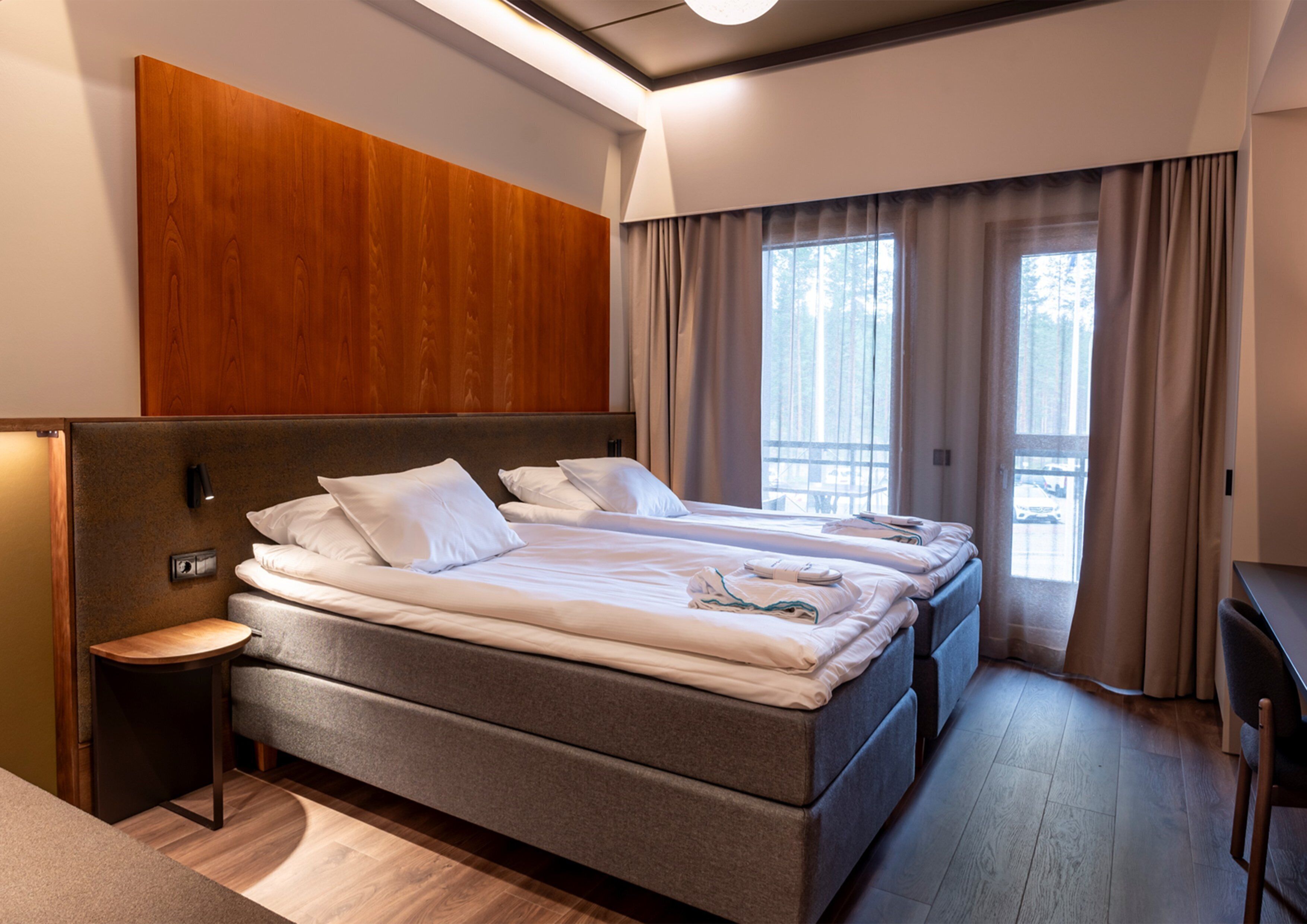 A modern hotel room with a large bed, a bedside table, and a view of the outdoors through the window.
