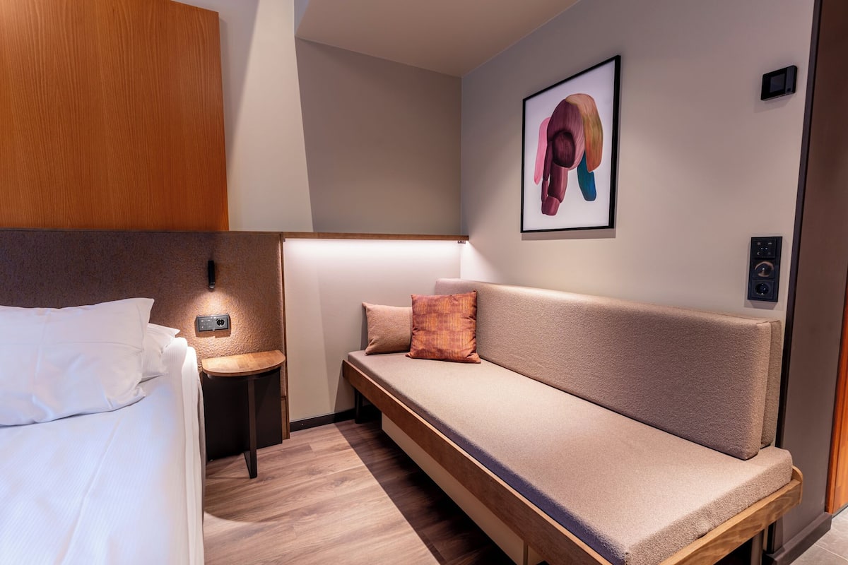 A modern hotel room with a bed, a sofa, a small side table, and a wall-mounted light.
