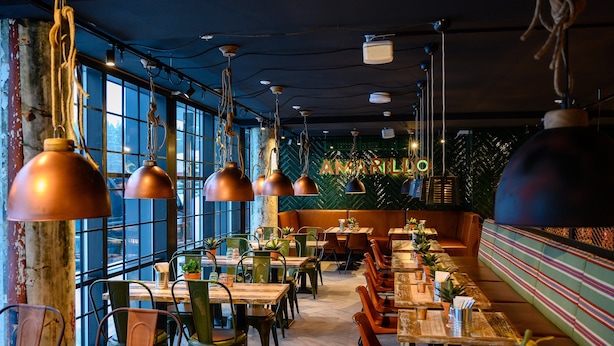Een restaurantinterieur met hoge plafonds, zichtbare ventilatiekanalen en grote ramen. Tafels en stoelen zijn klaar voor het diner.