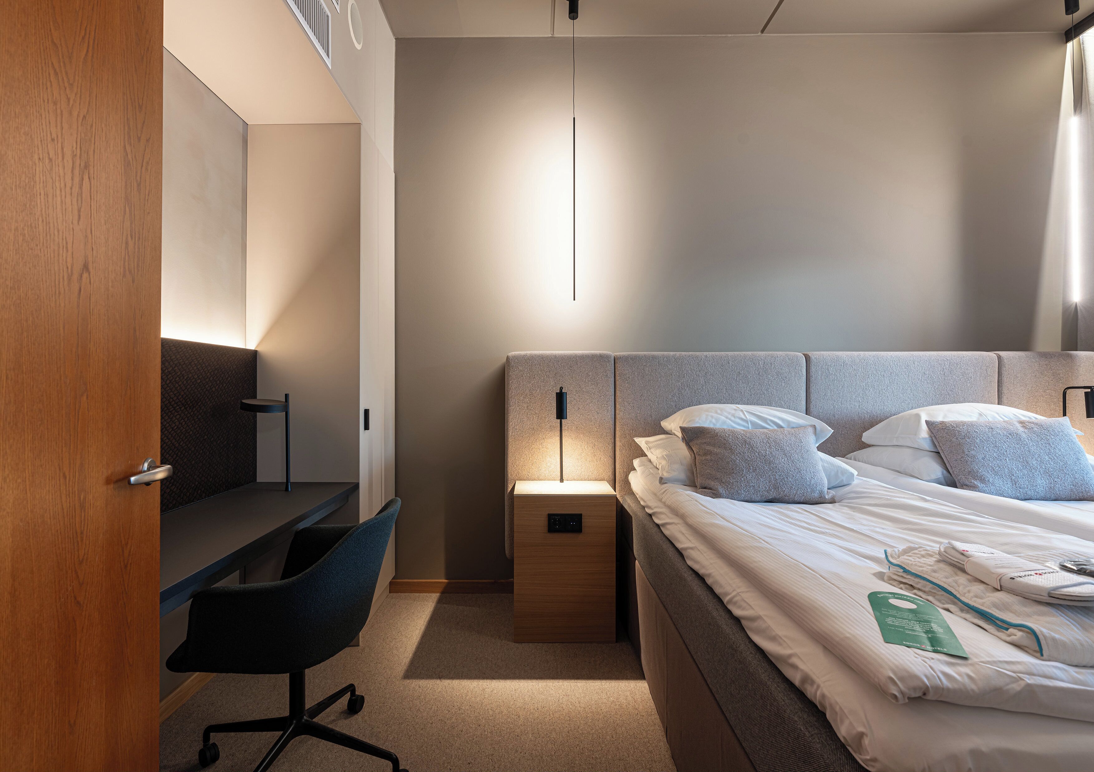Quarto de hotel moderno com escrivaninha de madeira, cama confortável com almofadas e mesa de cabeceira com abajur.