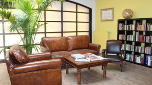 Quarto com sofá de couro marrom, mesa de centro de madeira com revistas, poltrona de couro preta, uma planta grande em vaso e uma estante cheia de livros.