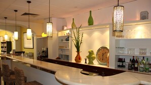 Uma área de bar com cadeiras de vime, balcão de mármore, luminárias pendentes, um relógio e prateleiras com garrafas e copos.