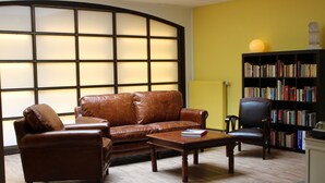 Uma sala de estar com um sofá de couro marrom, uma mesa de centro de madeira e uma poltrona preta. Uma estante cheia de livros está encostada à parede amarela.
