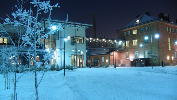 A snow-covered outdoor area with a building that has a modern facade and a brick structure. There are lights illuminating the area, and a person is visible in the distance. - Hotel Lasaretti (Oulu)