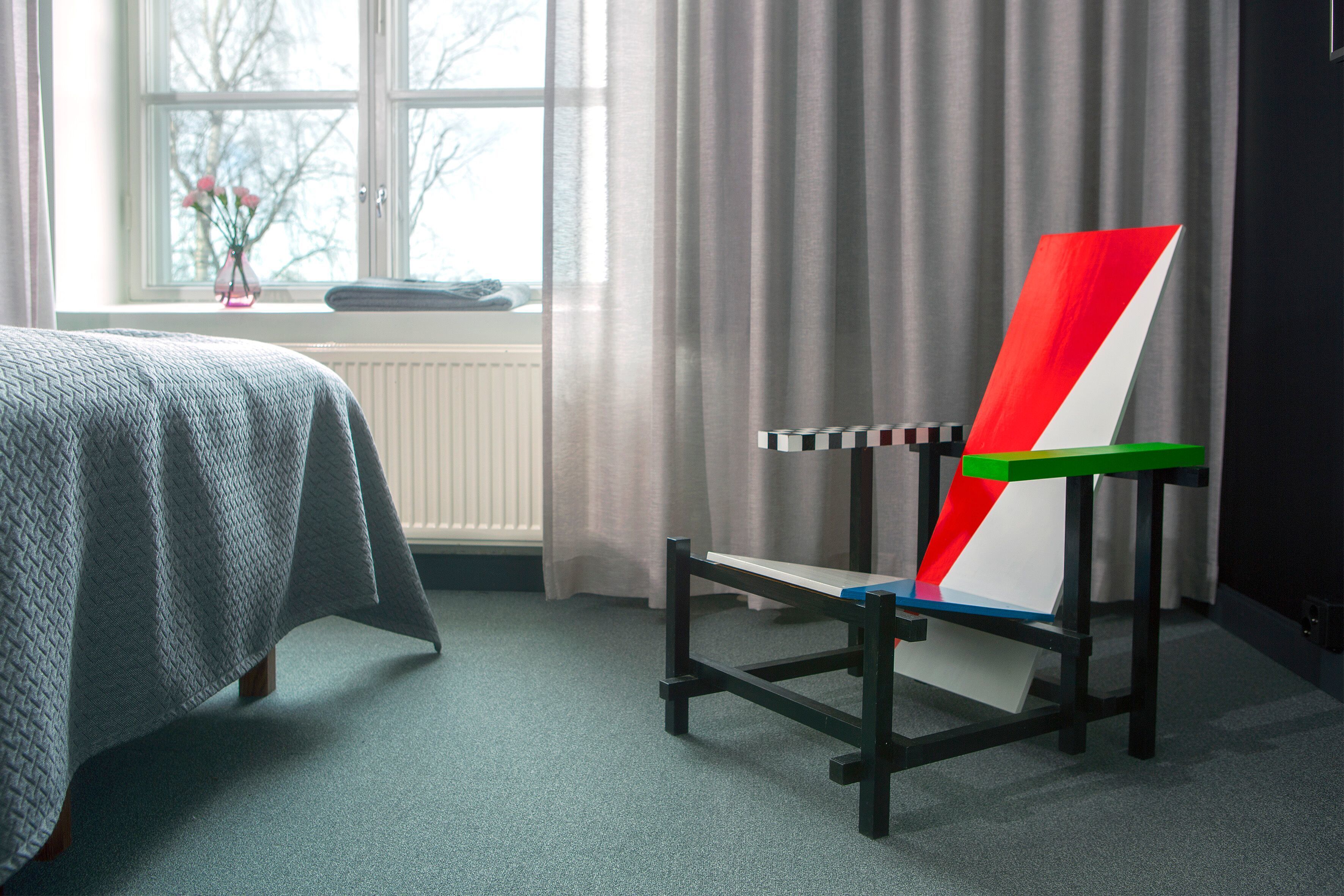 Une pièce avec un lit, une fenêtre avec des rideaux et une chaise dont le dossier est de couleur vive.
