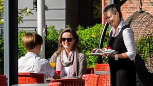 An outdoor cafe with red chairs and tables, a waiter serving food, and a brick building in the background. - Hotel Lasaretti (Oulu)