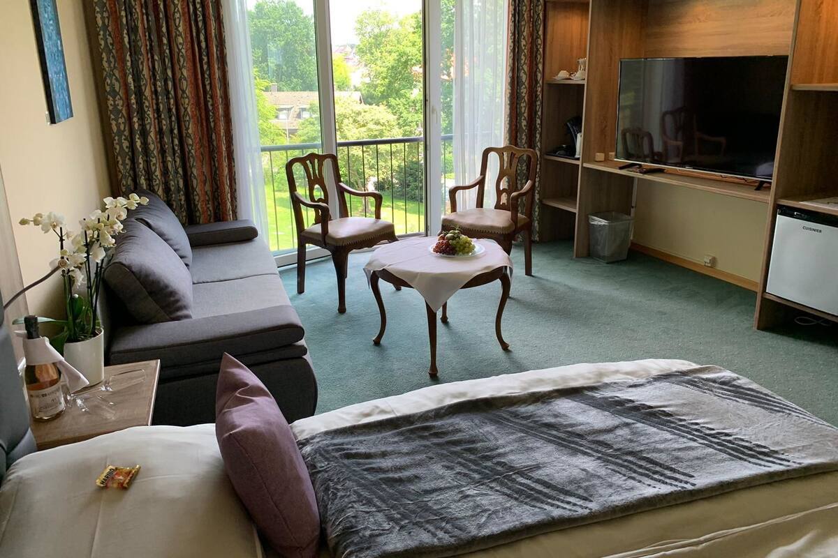 A hotel room with a sofa, two chairs, a small table with fruit, a TV, and a balcony with a view of greenery.