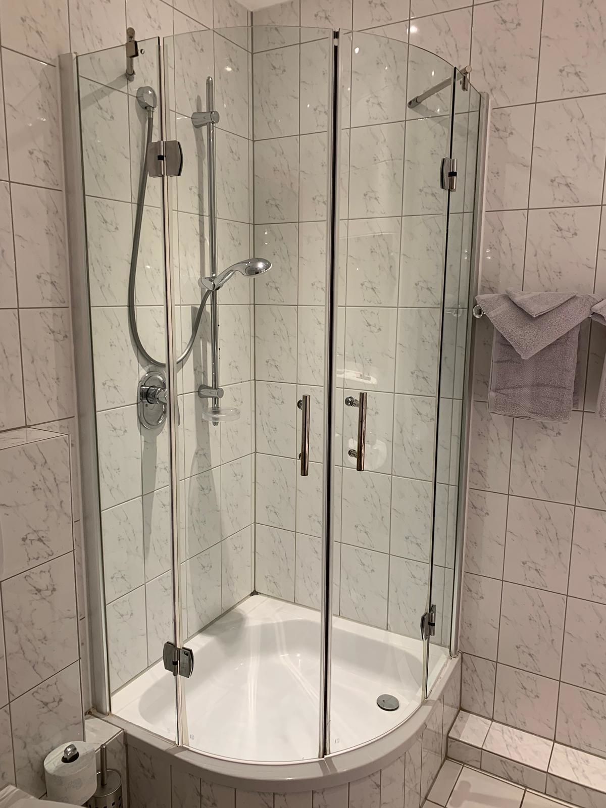 Ducha en esquina con puertas de vidrio, un lavamanos blanco y un perchero.