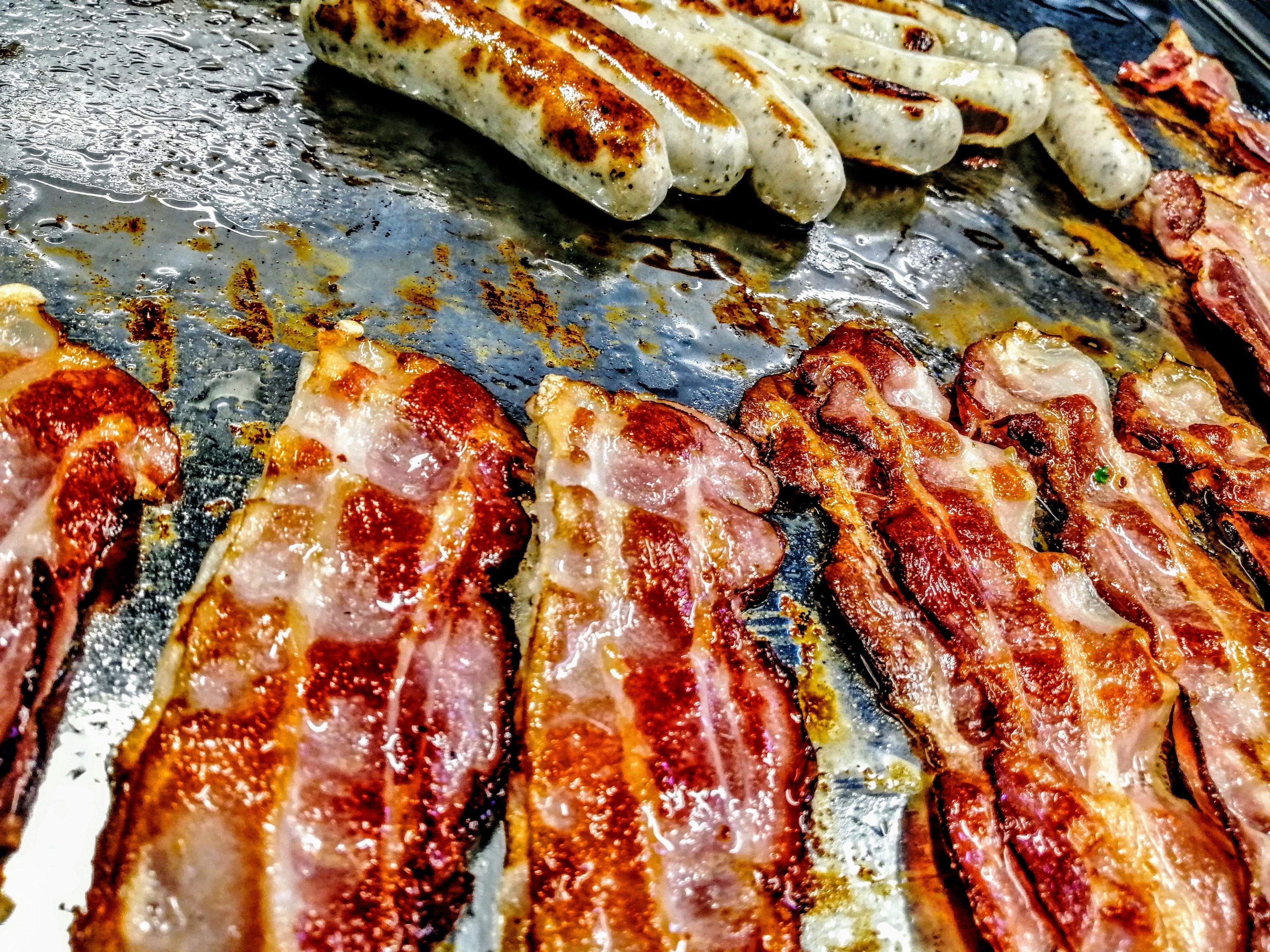 Primer plano de salchichas asadas y tocino sobre una superficie de cocción.